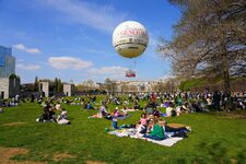 Una escena primaveral en el Parque André Citroën en París: los habitantes se reúnen para un picnic amigable sobre la hierba verde, bañados en la suave luz de abril. En el fondo, un globo aerostático (Ballon Generali) flota pacíficamente, añadiendo un toque de magia y aventura a este cuadro vivo de la vida parisina en la orilla izquierda.