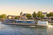 Un bateau vitré des Bateaux Parisiens navigue sur la Seine au coucher du soleil à Paris, avec les quais et l’Institut de France en arrière-plan.
