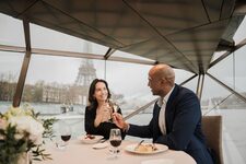 Un couple trinque avec des coupes de champagne à bord d’un bateau vitré naviguant sur la Seine, avec la Tour Eiffel visible en arrière-plan par temps gris.
