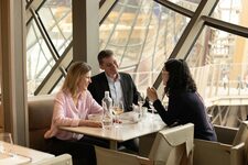 Trois personnes déjeunent à une table élégante dans la Madame Brasserie, au premier étage de la Tour Eiffel, avec la structure métallique visible à travers les grandes baies vitrées.