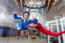 Two people in jumpsuits perform indoor skydiving at the iFLY simulator in Paris, floating weightlessly inside the glass tunnel.