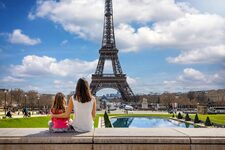 A woman and a young girl sit facing the Eiffel Tower from the Trocadéro gardens, enjoying a sunny outdoor day.