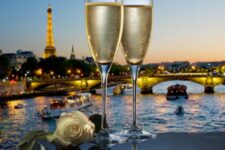 Two glasses of champagne on a table with a white rose, overlooking the illuminated Seine and Eiffel Tower at sunset.